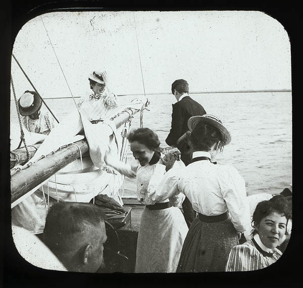"Yachting." View from the center of a boat, across the bow, with the sail down around the boom. Several women in Edwardian dress on different parts of the boat, while a man with his back to the camera faces the water, looking down. Photograph by T.S. and R.C. Stewart. Collection labeled "Medical School Donations," possibly from the collection of the Carpenter family. Penn Museum image #218711